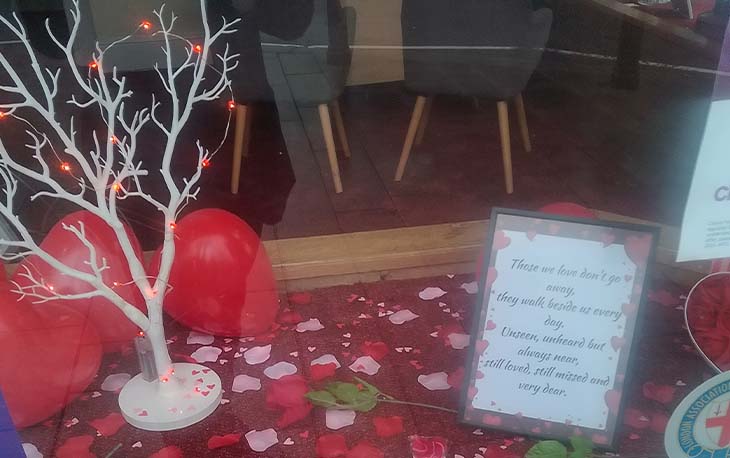 Picture of the front of a shop window with red balloon hearts, pink and white heart confetti and a small white tree with red fairy lights wrapped around.