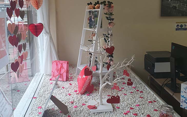 Picture of the inside of a display window showing a bunch or red and pink heart bunting. Also a white tree with hearts hanging from the branches.